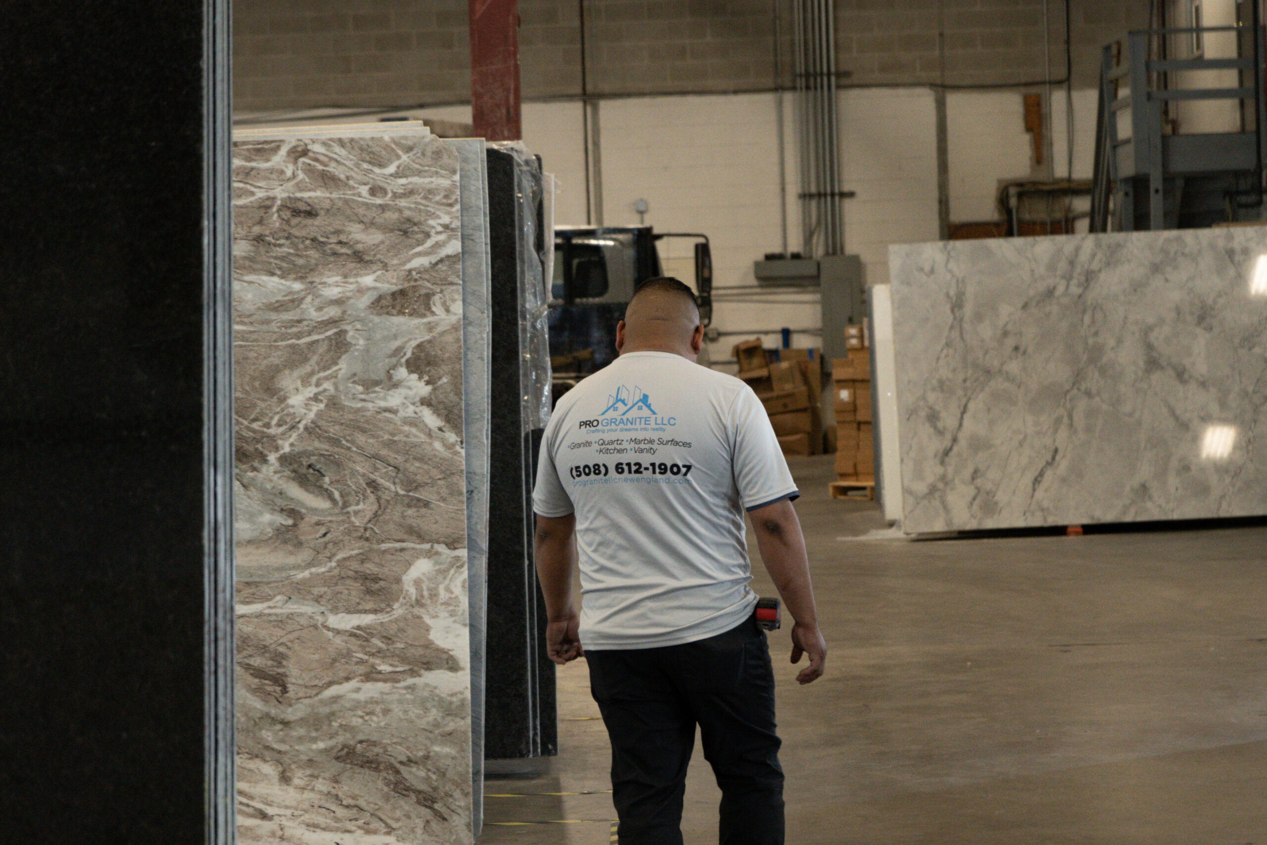 A Pro Granite LLC team member in a branded company shirt walking between granite and marble slabs stored upright in the Worcester fabrication warehouse