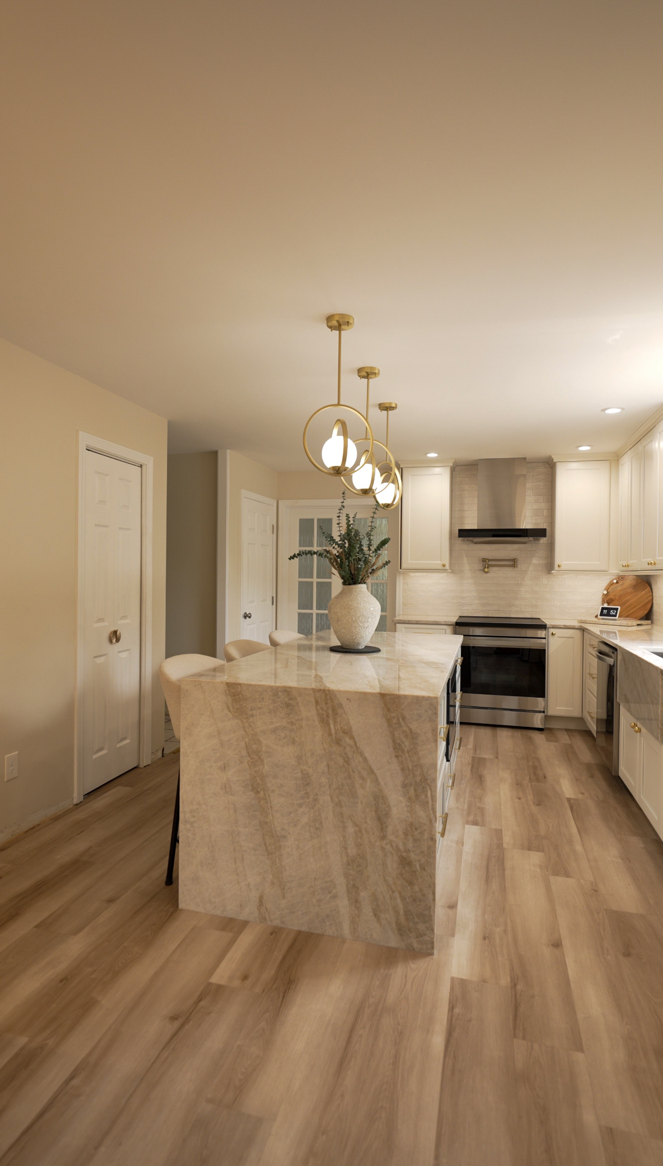 Kitchen with a warm beige quartzite waterfall island, brass ring pendant lights, cream shaker cabinetry, a stainless range and hood, and light wood flooring