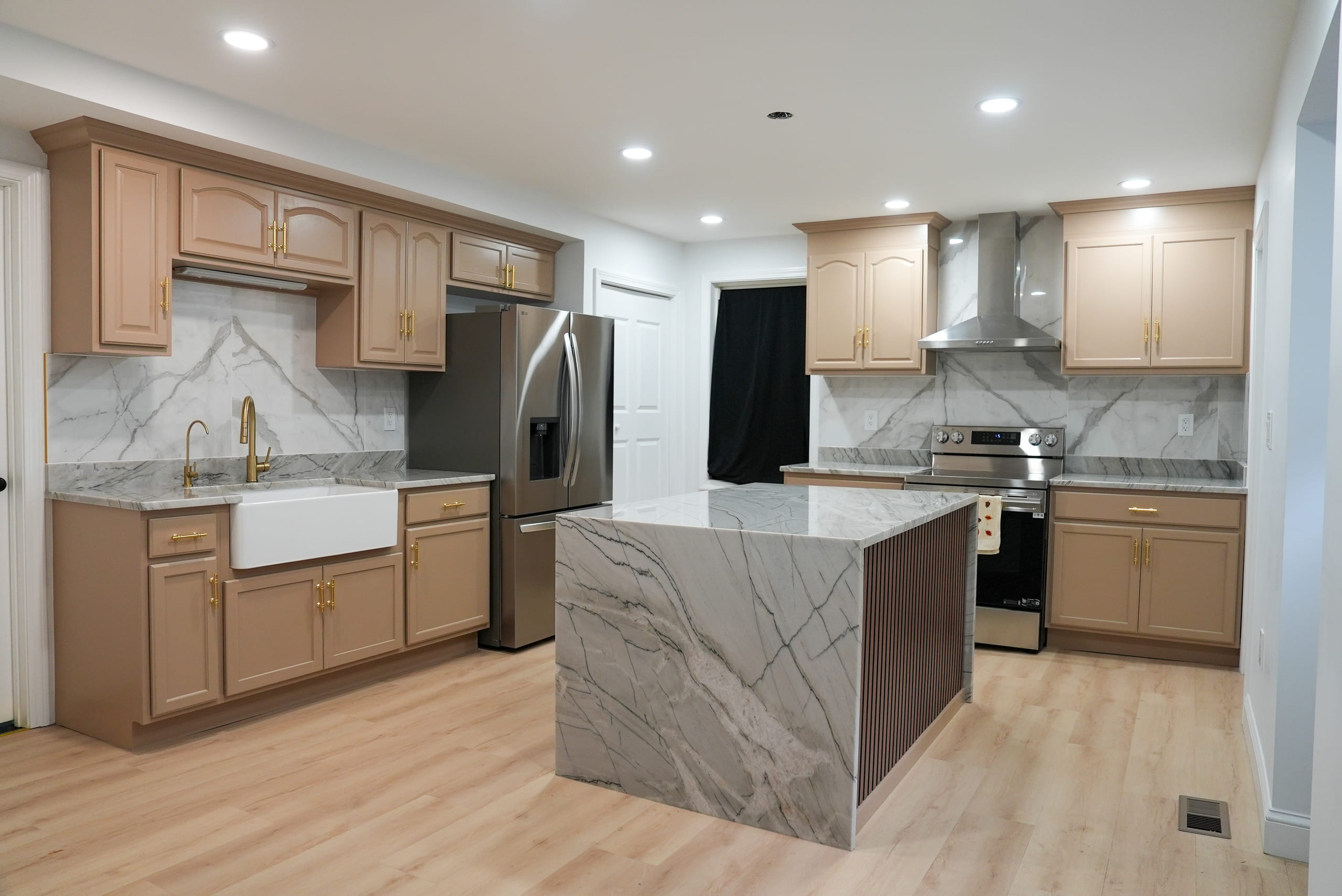 Kitchen in warm taupe raised-panel cabinetry with brushed gold hardware, a dramatic grey and white marbled island with a waterfall edge and fluted base, a farmhouse sink and a full-height stone backsplash