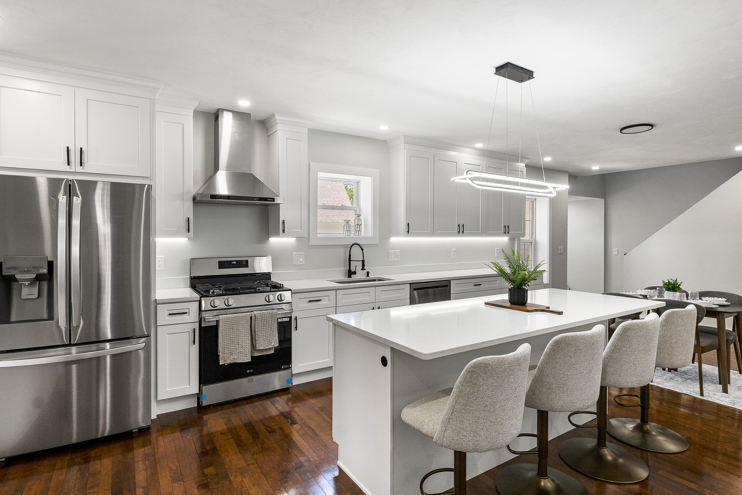 White shaker kitchen with a long white quartz island seating three, a stainless chimney hood over a gas range, a linear pendant light and dark hardwood flooring