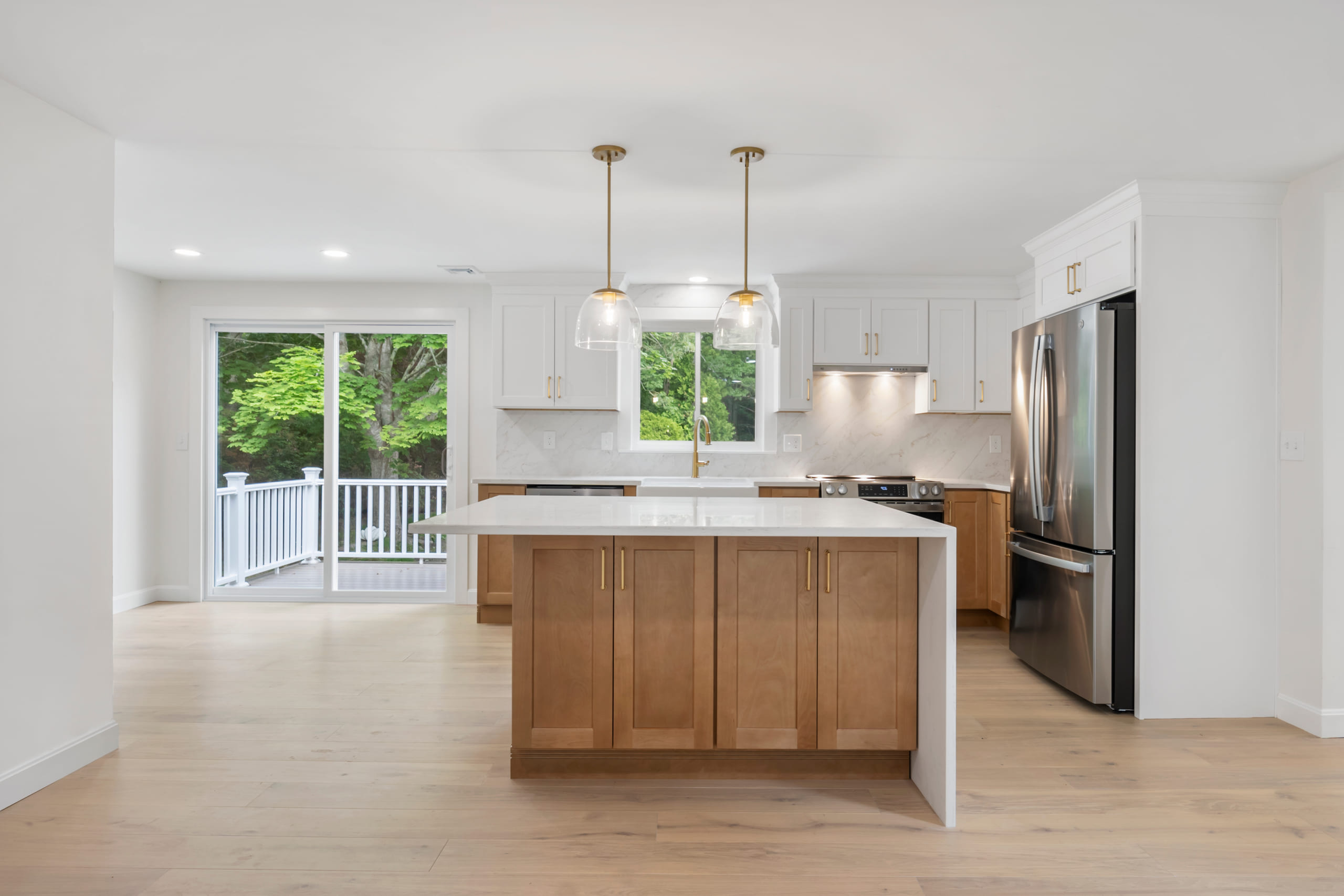A bright white kitchen with a quartz waterfall island over stained wood cabinetry, brass pendant lights and a white oak floor, fabricated and installed by Pro Granite