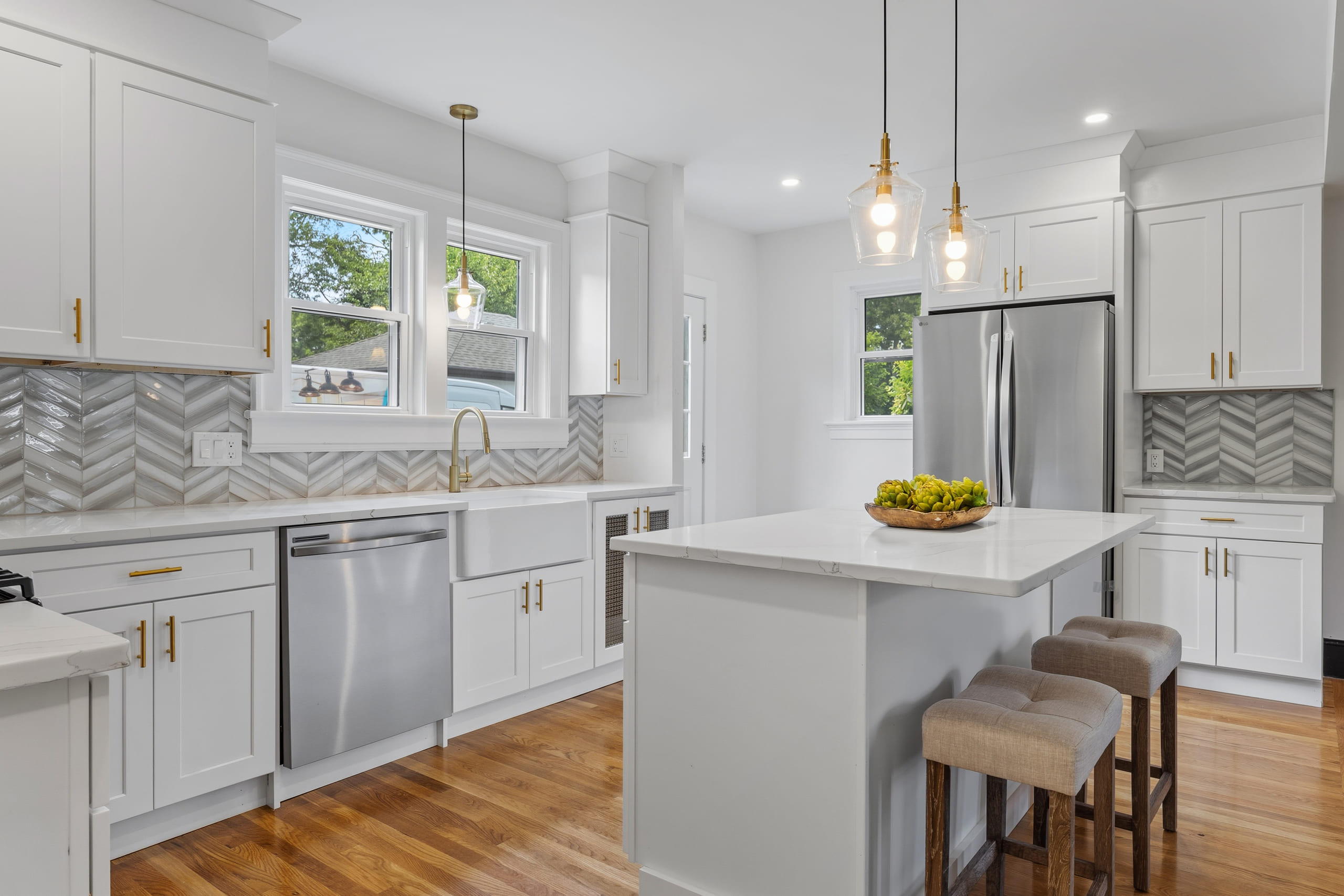 White shaker kitchen with brushed gold hardware and pendant lights, a grey chevron tile backsplash, a farmhouse sink under twin windows and a white quartz island with woven stools
