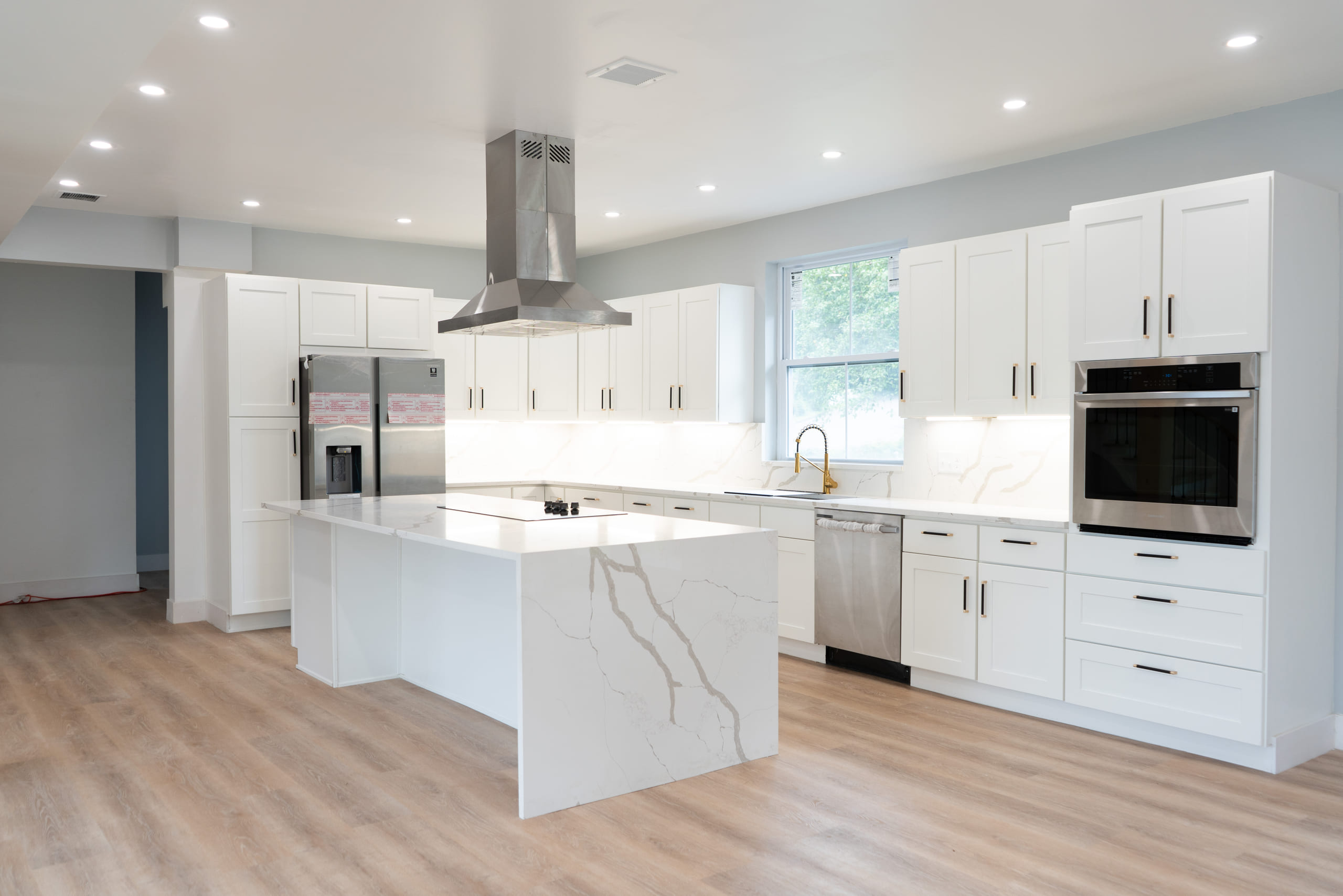 Bright white kitchen with a waterfall-edge quartz island veined in soft grey, a stainless steel island hood above it, a stacked wall oven and microwave, white shaker cabinetry and pale hardwood flooring