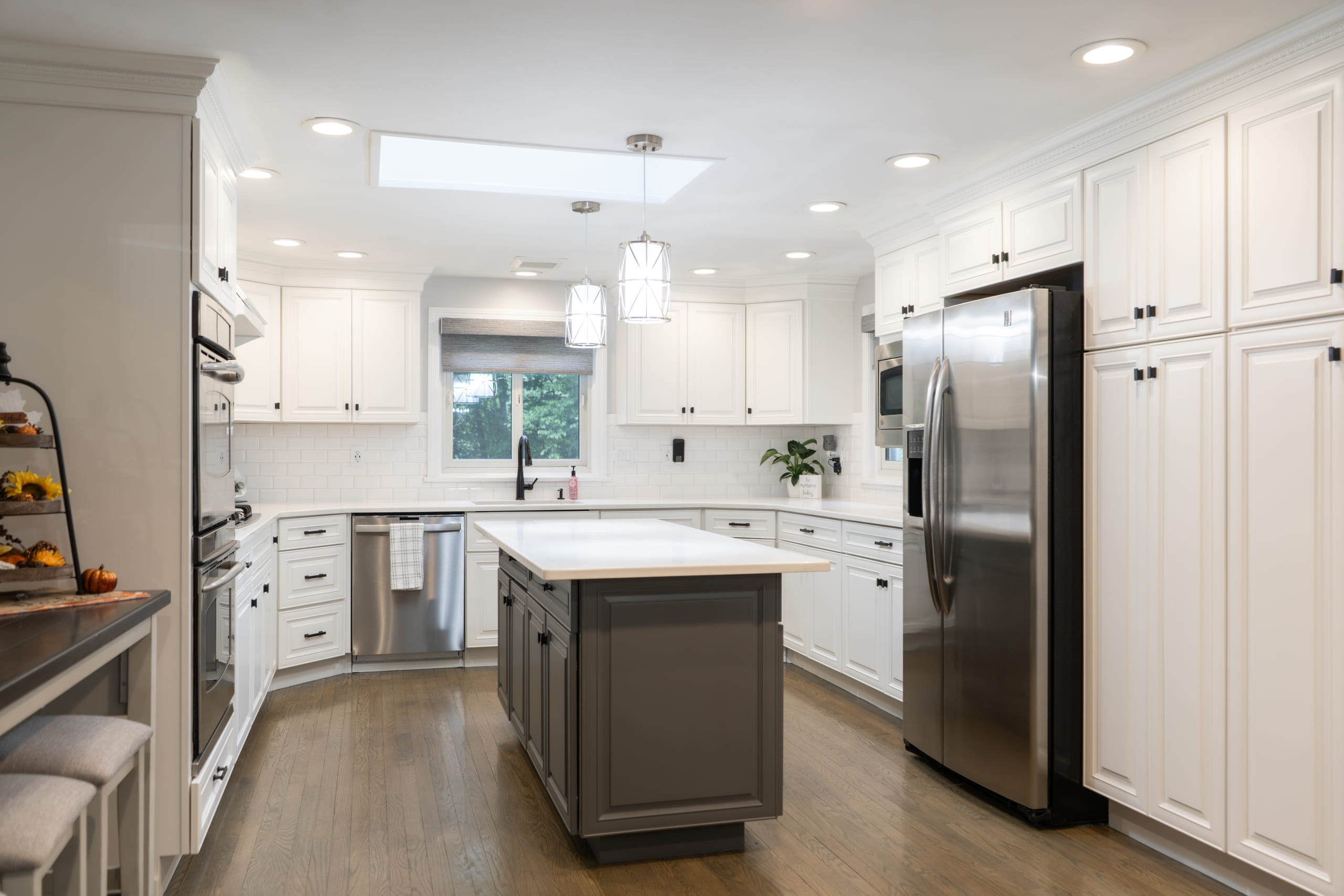 White kitchen lit by a skylight with a grey island under a glass lantern pendant, a wood-topped breakfast bar, double wall ovens and warm oak flooring