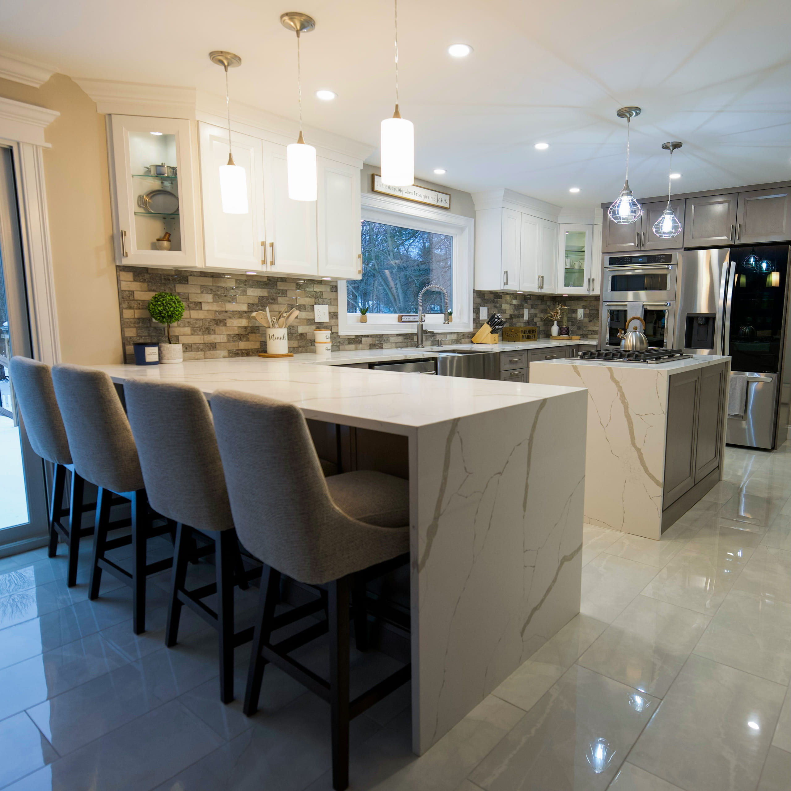 Kitchen peninsula topped in white quartz with grey veining and a waterfall edge, four grey upholstered stools along it, glass pendant lights above and a mosaic tile backsplash behind grey and white cabinetry